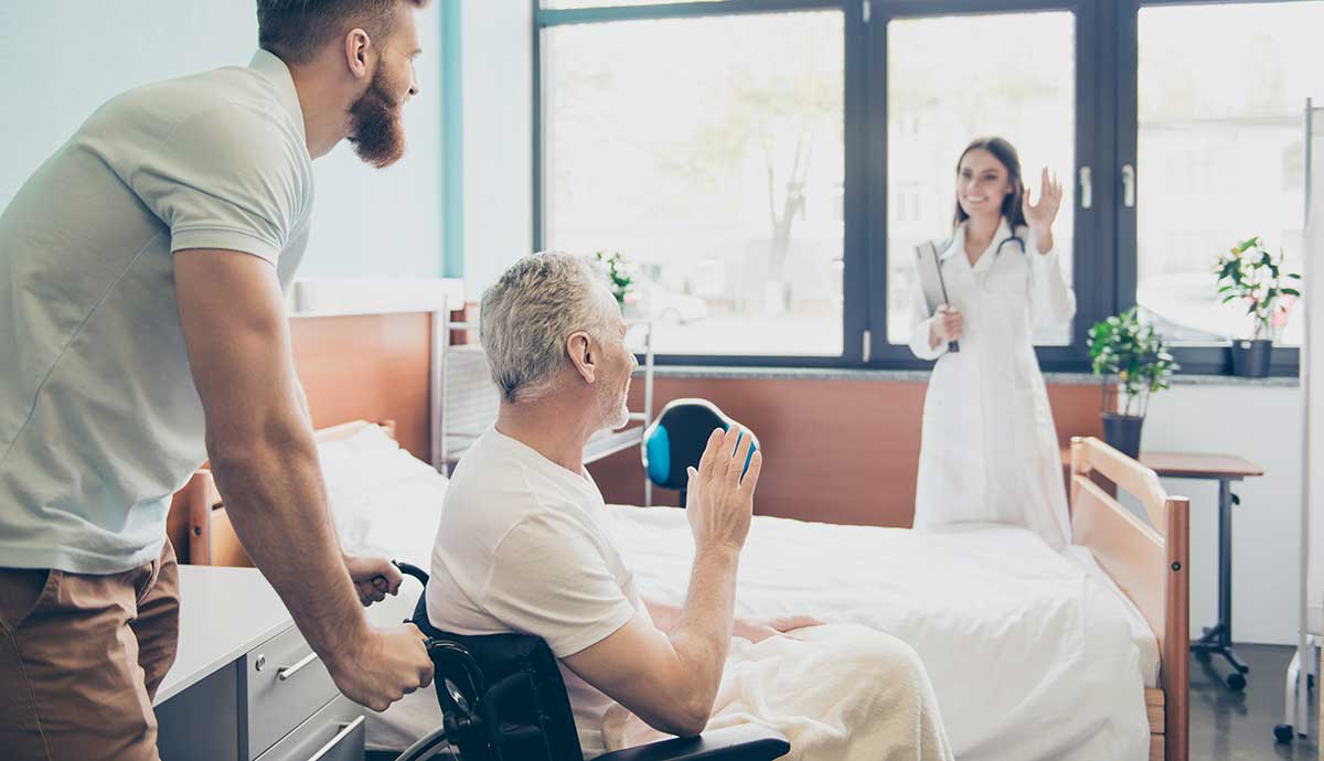 A smiling doctor waves at an older male patient in a wheelchair, who is being pushed by a caregiver in a brightly lit hospital or rehabilitation room.